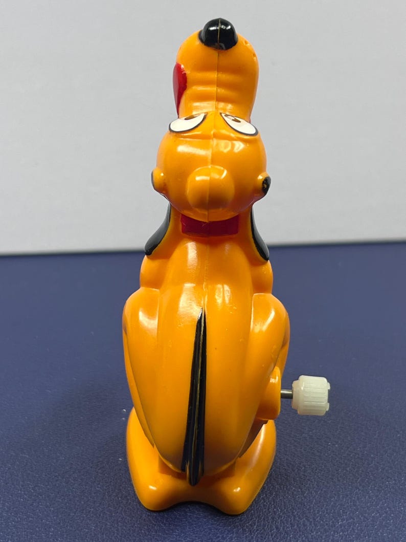 May include: A vintage Pluto wind-up toy. The toy is primarily orange with black ears and a black stripe down its back. It has a red bow tie and a white crank on the side. The toy is a collectible item.