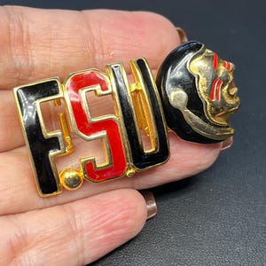 May include: A gold-tone metal brooch featuring the letters FSU in black and red enamel, accompanied by a stylized figure of a person wearing a black headdress with red facial markings. The design is finished with a polished gold-tone metal border.