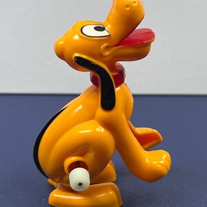 May include: A plastic toy figure of Pluto, a yellow dog with black ears and a black nose. The dog has a red collar and a red tongue. The toy is sitting on its haunches with its mouth open.