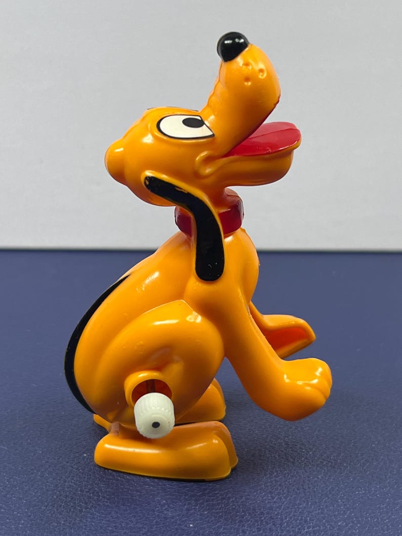 May include: A plastic toy figure of Pluto, a yellow dog with black ears and a black nose. The dog has a red collar and a red tongue. The toy is sitting on its haunches with its mouth open.