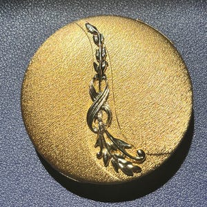 Può includere: Un compatto rotondo color oro con superficie strutturata. Un design floreale stilizzato color argento adorna la parte superiore. Il compatto è posto su uno sfondo blu scuro.