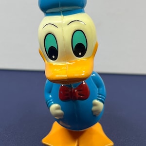 May include: A plastic Donald Duck figurine. The figure has a blue hat and shirt, a yellow face and feet, and a red bow tie. The eyes are large and green. The figurine is standing upright.