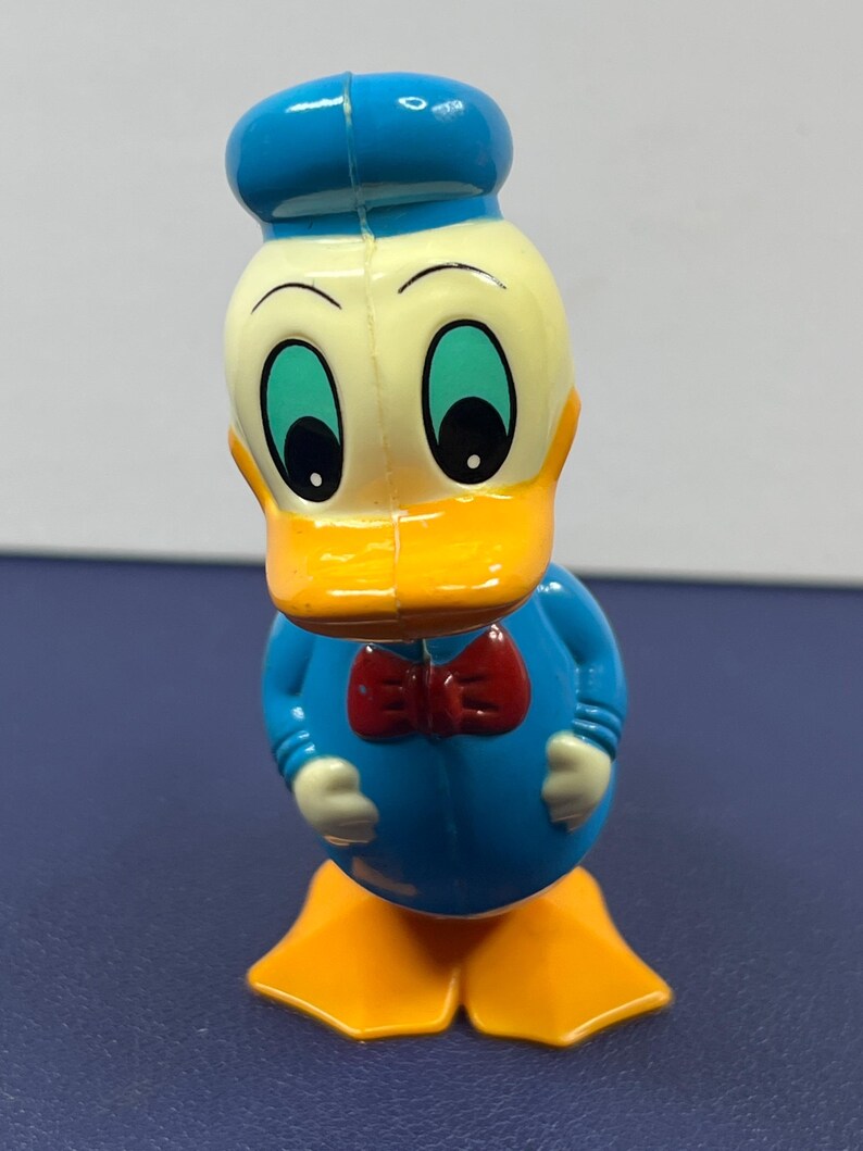 May include: A plastic Donald Duck figurine. The figure has a blue hat and shirt, a yellow face and feet, and a red bow tie. The eyes are large and green. The figurine is standing upright.