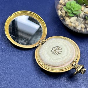 May include: Open, antique gold-toned compact with a mirror and powder. The compact is round with a hinged lid. The powder has a light pink base and a white powder puff with an ornate initial design. The compact is on a blue surface.