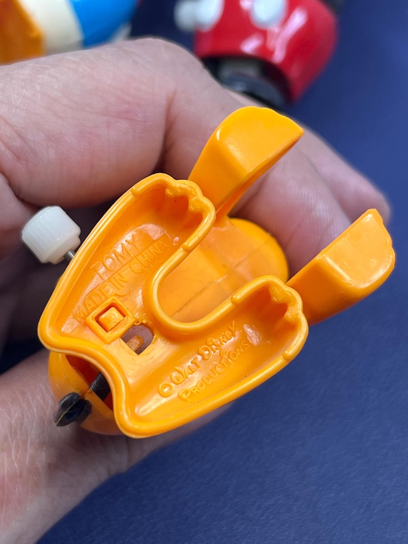 May include: Close-up of an orange plastic toy with a wind-up mechanism. The toy has a wing-like design and the text "TOMY MADE IN CHINA" is visible. The toy is a vintage Disney collectible.