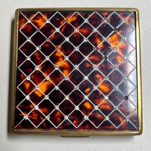 May include: A vintage tortoiseshell compact with a gold frame and a diamond pattern. The compact is decorated with small, clear rhinestones.