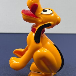 May include: A plastic figurine of Pluto, a cartoon dog. The figurine is primarily orange with black, red, and white accents. Pluto is depicted in a seated position with his tongue sticking out. The figurine has a red collar and black ears.