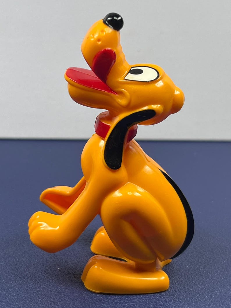 May include: A plastic figurine of Pluto, a cartoon dog. The figurine is primarily orange with black, red, and white accents. Pluto is depicted in a seated position with his tongue sticking out. The figurine has a red collar and black ears.