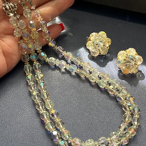 Puede incluir: Un conjunto de joyas vintage con un collar de varias hebras y pendientes a juego. El collar y los pendientes están hechos de cuentas transparentes y facetadas con cualidades iridiscentes. El collar tiene un cierre plateado.