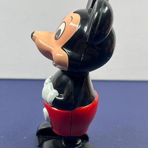 May include: A vintage Mickey Mouse figurine. The figure has a black head and ears, a tan face, and a red pair of shorts. The figure's hands are white, and the feet are black. The figurine is a classic Disney collectible.