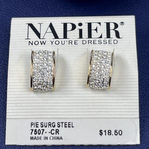 Puede incluir: Un par de pendientes de aro dorados incrustados con cristales transparentes. Los pendientes se exhiben en una tarjeta blanca con el logotipo de Napier y el texto "NOW YOU'RE DRESSED". La tarjeta también incluye información del producto.