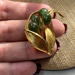Puede incluir: Un broche dorado con un grupo de piedras de jade verde engastadas en un diseño de hojas.