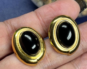 May include: A pair of gold-toned oval earrings with a black, glossy center stone. The earrings have a simple, elegant design, with a smooth, polished finish. The earrings are held in a person's hand.