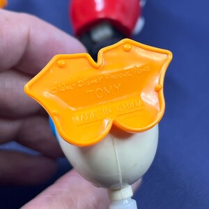 May include: Close-up of a vintage Donald Duck toy figure. The figure has a white head, blue bow tie, and an orange base with the text "Walt Disney Productions" and "TOMY". The toy is made in China.