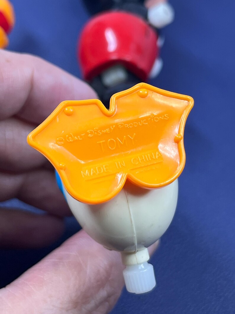 May include: Close-up of a vintage Donald Duck toy figure. The figure has a white head, blue bow tie, and an orange base with the text "Walt Disney Productions" and "TOMY". The toy is made in China.