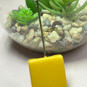 Può includere: Un ciondolo quadrato in vetro giallo con una parte superiore in metallo argentato attaccata a un filo metallico sottile.