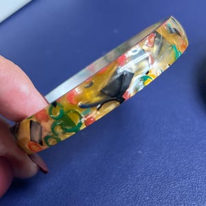 Op de afbeelding: Een kleurrijke armband met een mozaïekontwerp. De armband heeft een mix van kleuren, waaronder geel, zwart, rood en groen. De armband heeft een solide, afgeronde vorm.