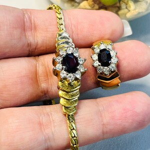 May include: A gold-coloured bracelet and ring set. The bracelet has a chain design with a central floral-shaped cluster of a dark red stone surrounded by clear crystals. The ring has a similar design, with a dark red stone and clear crystals.