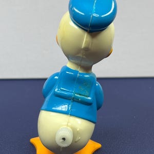 May include: A vintage Donald Duck toy figurine, viewed from the back. The toy features a blue sailor hat and jacket, a cream-colored body, and orange feet. A wind-up mechanism is visible on the back.