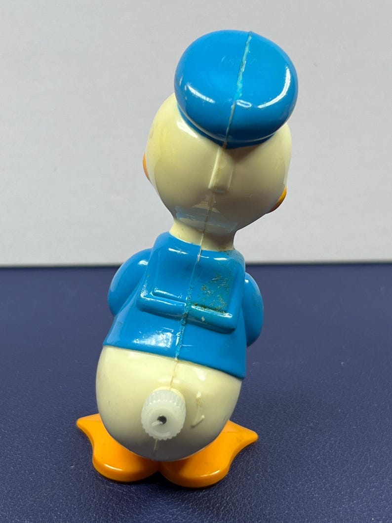 May include: A vintage Donald Duck toy figurine, viewed from the back. The toy features a blue sailor hat and jacket, a cream-colored body, and orange feet. A wind-up mechanism is visible on the back.