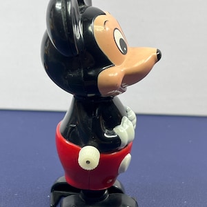 May include: A vintage Mickey Mouse figurine. The figure has a black head and body, a tan face, and a red pair of shorts. The figure is standing on a black base. The figurine is a classic Disney collectible.