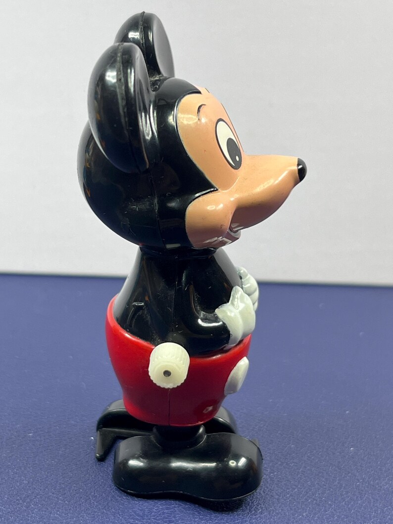 May include: A vintage Mickey Mouse figurine. The figure has a black head and body, a tan face, and a red pair of shorts. The figure is standing on a black base. The figurine is a classic Disney collectible.