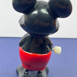 May include: A vintage Mickey Mouse wind-up toy. The toy features a black head with large ears, a black body, red shorts, and black shoes. A white wind-up key is on the side. The toy is made of plastic.