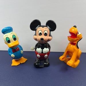 May include: Three vintage Disney character wind-up toys: Donald Duck in blue, Mickey Mouse in black and red, and Pluto in orange. The toys are plastic and have a classic design, perfect for collectors or fans of Disney.