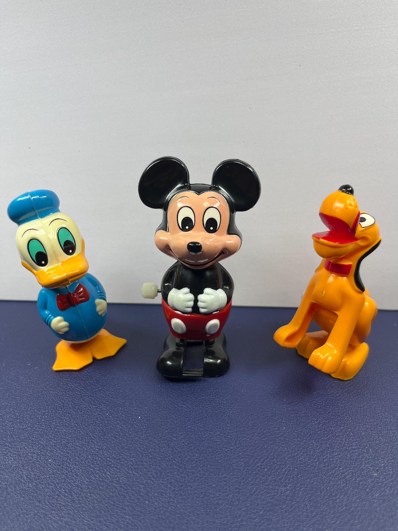 May include: Three vintage Disney character wind-up toys: Donald Duck in blue, Mickey Mouse in black and red, and Pluto in orange. The toys are plastic and have a classic design, perfect for collectors or fans of Disney.