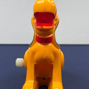 May include: A vintage wind-up toy dog in bright orange, red, black, and white. The toy dog has a red collar and black ears. The wind-up key is on the left side. The toy is a collectible item.