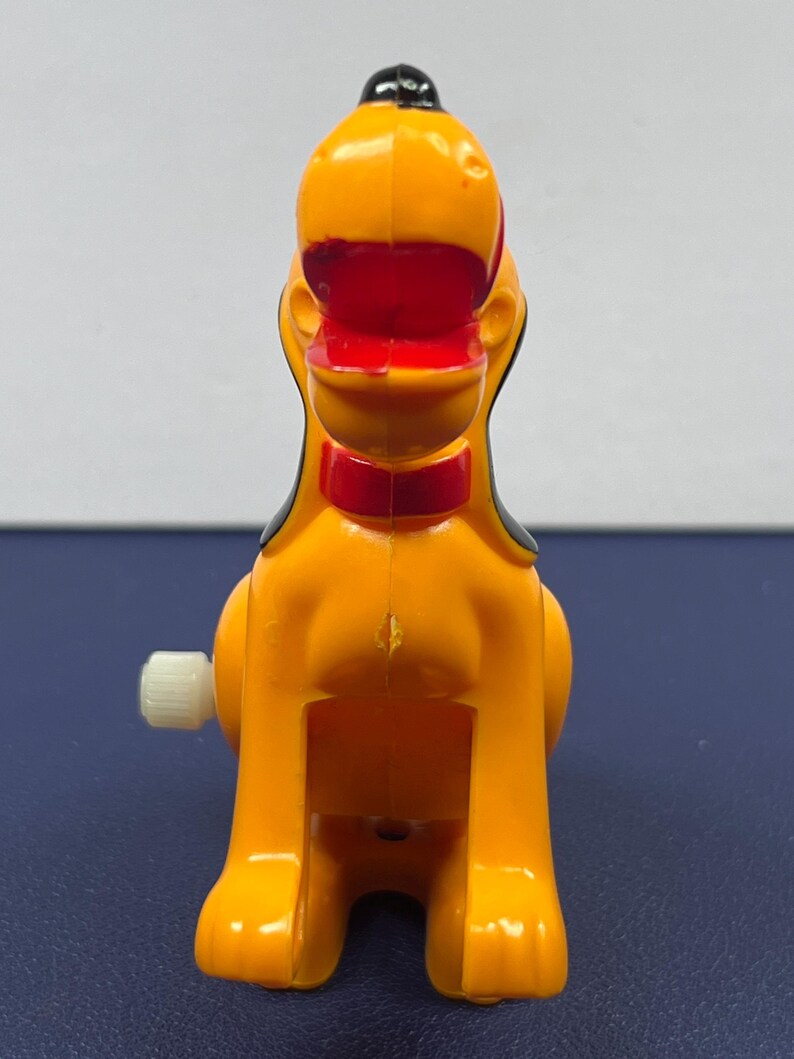 May include: A vintage wind-up toy dog in bright orange, red, black, and white. The toy dog has a red collar and black ears. The wind-up key is on the left side. The toy is a collectible item.