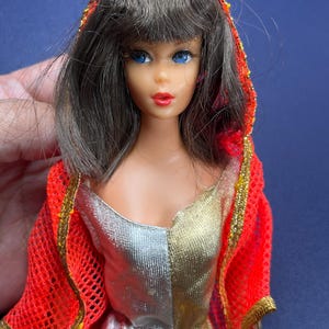 Könnte beinhalten: Eine Vintage-Puppe mit blauen Augen, roten Lippen und braunem Haar. Die Puppe trägt einen gold-silbernen Overall und einen roten Netzschal mit goldenen Verzierungen. Das Gesicht der Puppe ist detailliert mit bemalten Merkmalen.