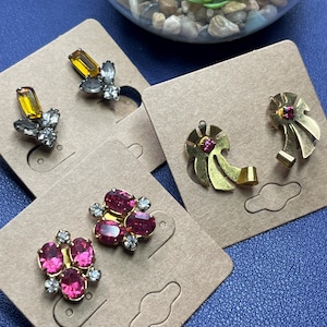 May include: A selection of vintage earrings presented on tan card. The earrings showcase diverse designs, including gold-toned ribbon shapes with pink gems, and clusters of pink and clear rhinestones. A glass terrarium with plants is in the background.