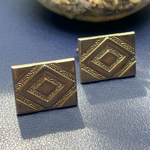 May include: A pair of gold-toned square cufflinks. Each cufflink features a raised diamond pattern with a textured border. The cufflinks are displayed on a blue surface, with a blurred background.