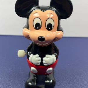 May include: A vintage Mickey Mouse wind-up toy. The toy features a black body, large black ears, and a red shorts. The face is light pink with large eyes and a smiling expression. The toy has white gloves and black shoes.