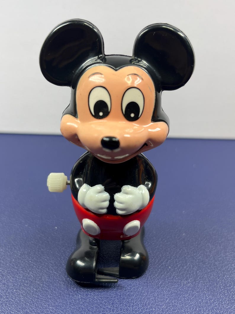 May include: A vintage Mickey Mouse wind-up toy. The toy features a black body, large black ears, and a red shorts. The face is light pink with large eyes and a smiling expression. The toy has white gloves and black shoes.