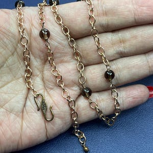 Può includere: Una collana color oro con un design a catena, con perline rotonde marrone scuro. La collana è esposta su una mano, mostrando gli intricati dettagli del gioiello. La chiusura è visibile, aggiungendo al design generale.