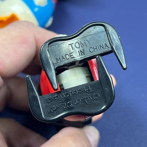 May include: Close-up of the underside of a vintage toy figure. The black plastic base has the text "TOMY MADE IN CHINA" and "&copy; Walt Disney Productions". The figure has red, black, and white details.