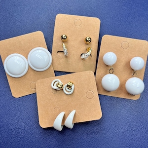 May include: A selection of white earrings displayed on brown card. Styles include large button earrings, gold stud earrings with white feather details, white ball drop earrings, gold and white hoop earrings, and white half-moon earrings. The background is blue.
