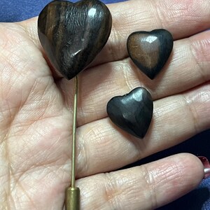 Peut inclure: Un ensemble de trois accessoires en bois en forme de cœur. L'un est une épingle avec une tige dorée et un cœur brun foncé. Les deux autres sont de petits boutons ou clous en forme de cœur brun foncé. Le grain du bois est visible.