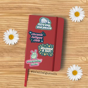 K&ouml;nnte beinhalten: Ein rotes Notizbuch mit Aufklebern mit den Aufschriften "flare day, dnd please", "chronic fatigue club", "disabled & hot!" und "everything hurts". Ein rotes Band dient als Lesezeichen. G&auml;nsebl&uuml;mchen umgeben das Notizbuch.