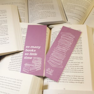 Peut inclure: Deux marque-pages roses avec des contours blancs d'une pile de livres. Le texte sur les marque-pages est "so many books so little time". Le marque-page de droite a également le texte "de luz design studio".