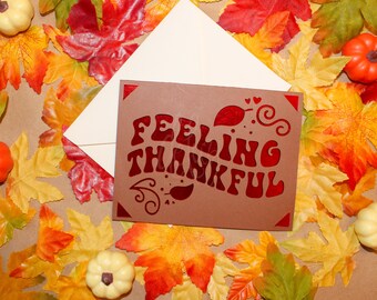 May include: A brown card with the words "Feeling Thankful" in red letters, surrounded by fall leaves and small pumpkins.
