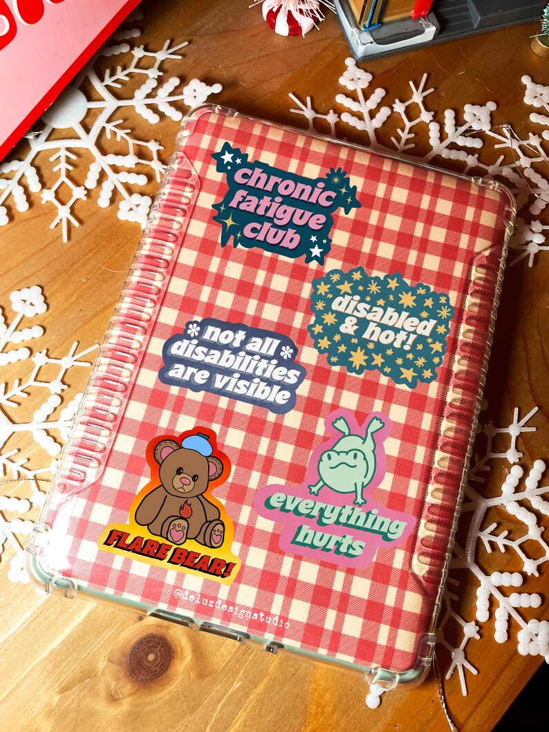 K&ouml;nnte beinhalten: Ein Notizbuch mit transparentem Kunststoffcover und rot-wei&szlig;em Karomuster. Es ist mit Aufklebern mit Texten wie "Chronic Fatigue Club", "Disabled & Hot!" und "Everything Hurts" versehen. Das Notizbuch hat auch einen B&auml;renaufkleber mit der Aufschrift "Flare Bear!".