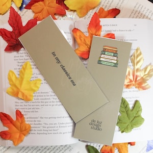 Puede incluir: Dos marcapáginas rectangulares beige. Uno con el texto "in my classics era", el otro con una pila de libros y "de luz design studio". Están sobre un libro abierto, con hojas de otoño.