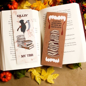Puede incluir: Dos marcapáginas de papel marrón con ilustraciones. El primer marcapáginas presenta un cuervo negro con una guadaña y una pila de libros con el texto "KILLIN' MY TBR". El segundo marcapáginas presenta una pila de libros con una boca de vampiro con el texto "A LITTLE SOMETHING TO SINK MY TEETH INTO".