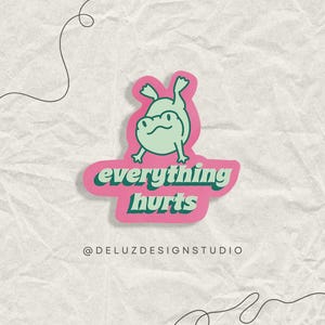 May include: A sticker featuring a light green frog with a pink background. The text "everything hurts" is written in a teal font. The sticker has a distressed paper background and the text "@DELUZDESIGNSTUDIO" is at the bottom.