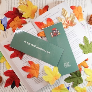 May include: Two green bookmarks with text that reads "in my dark academia era" and "de luz design studio". The bookmark on the left features a stack of books and a cat. The bookmarks are on a book with fall leaves scattered around.