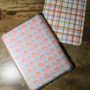 May include: A rectangular clear protective case featuring a repeating pattern of overlapping pink, orange, and light blue fan-shaped scales, positioned next to a rectangular card with a white, orange, pink, and green plaid grid pattern.