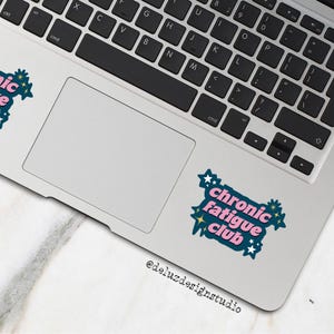 May include: A laptop with a keyboard and trackpad, featuring a sticker that reads "Chronic Fatigue Club" in pink and teal, surrounded by stars and floral accents. The sticker is on the trackpad.